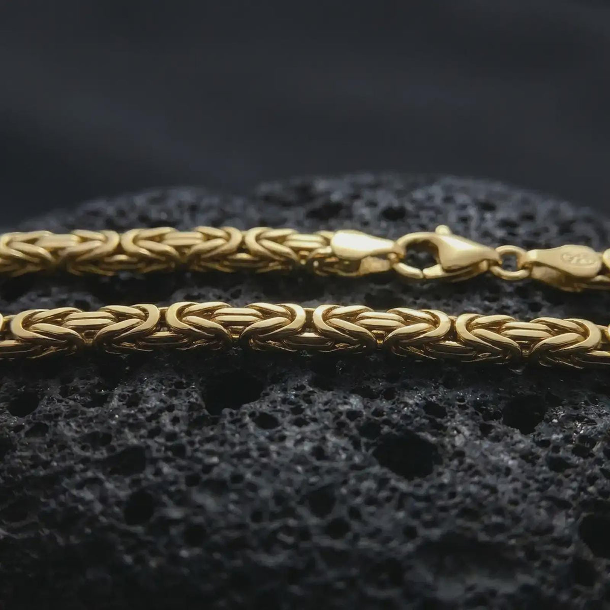 A gold plated 4mm square Byzantine chain necklace lies across rugged volcanic rock, showcasing its interwoven link design and strong masculine profile in a luxury macro close-up.