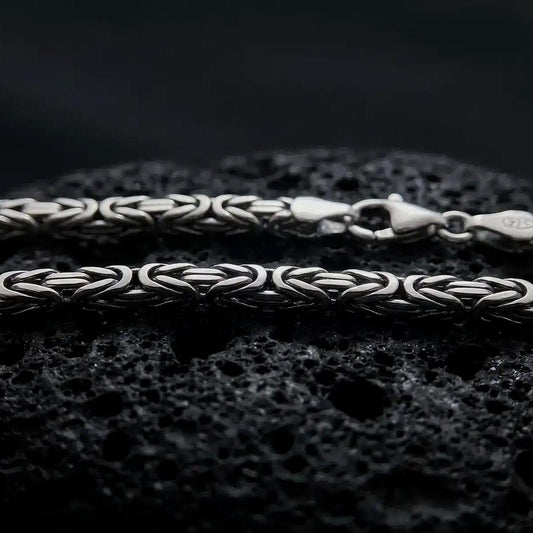 An oxidized 4mm square Byzantine chain necklace rests on dark volcanic rock, highlighting its tightly woven interlocking links and bold sterling silver structure in a macro studio close-up.