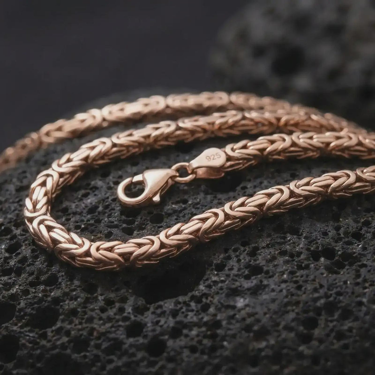 The rose gold plated 5mm square Byzantine necklace is arranged on black volcanic rock, emphasizing the structured link construction, flexible chain movement, and refined statement design in a detailed macro image.