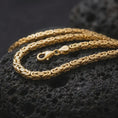 Load image into Gallery viewer, A gold plated 5mm square Byzantine chain necklace lies across rugged volcanic rock, showcasing its intricate interwoven link pattern and bold masculine chain profile in a luxury macro close-up.
