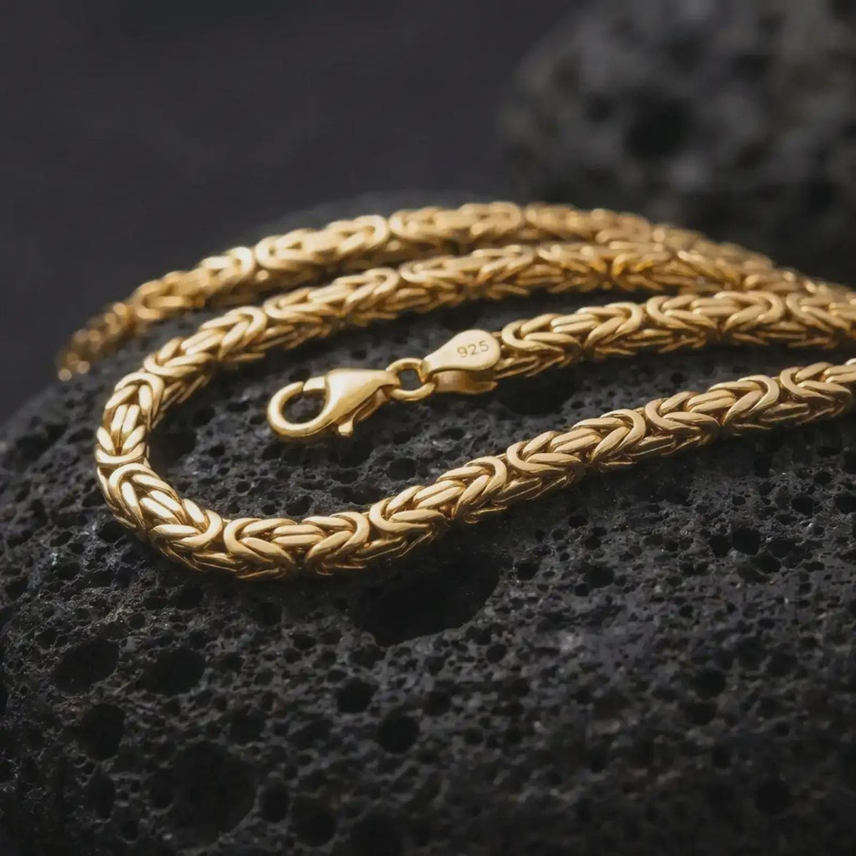A gold plated 5mm square Byzantine chain necklace lies across rugged volcanic rock, showcasing its intricate interwoven link pattern and bold masculine chain profile in a luxury macro close-up.