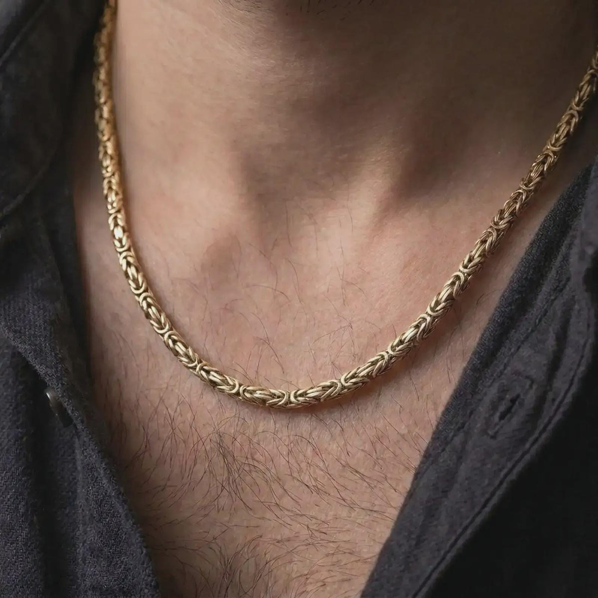 A gold plated 5mm square Byzantine chain necklace is worn on a man’s neck with dark clothing, highlighting the geometric woven links and bold statement appeal in a refined close-up.