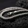 Load image into Gallery viewer, A 5mm square Byzantine chain necklace with an oxidized finish rests on dark volcanic rock, showing its tightly woven interlocking links and dimensional sterling silver structure in a macro studio close-up.
