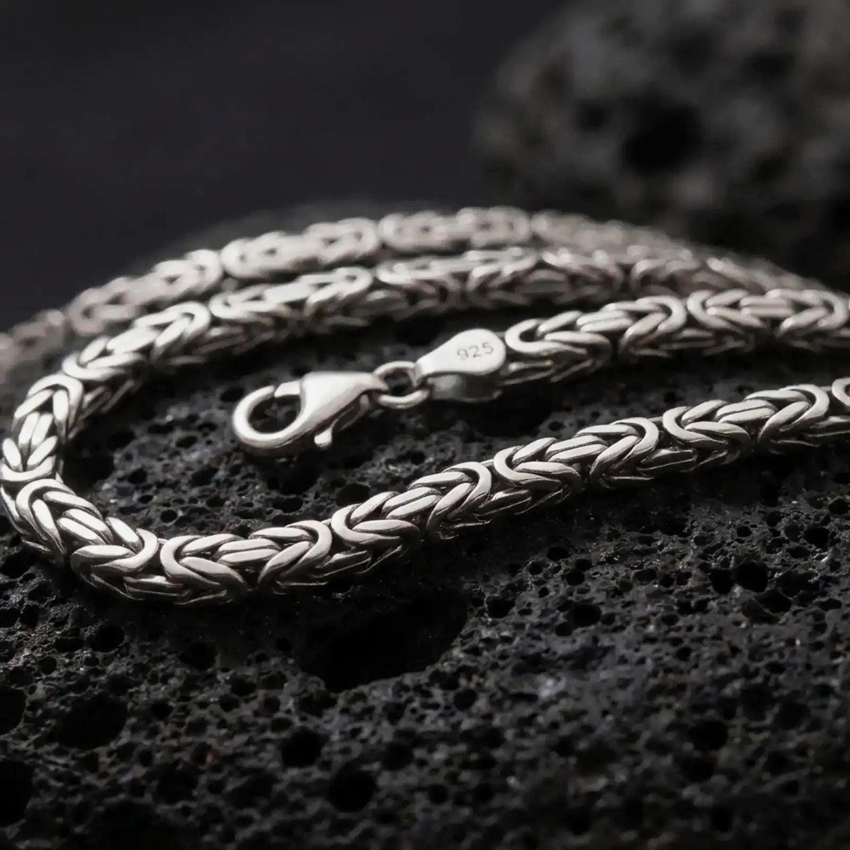 A 5mm square Byzantine chain necklace with an oxidized finish rests on dark volcanic rock, showing its tightly woven interlocking links and dimensional sterling silver structure in a macro studio close-up.