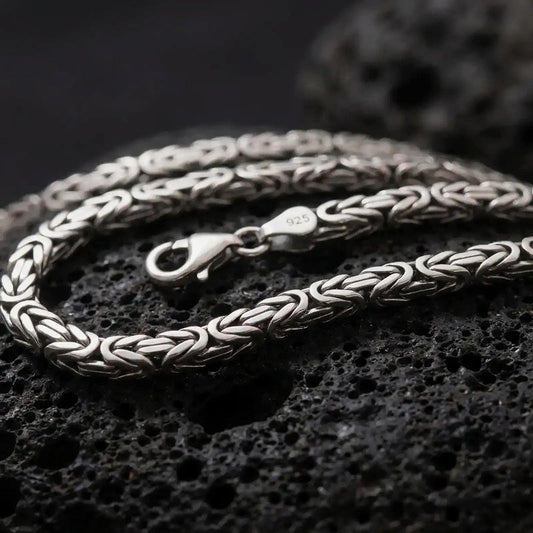 A 5mm square Byzantine chain necklace with an oxidized finish rests on dark volcanic rock, showing its tightly woven interlocking links and dimensional sterling silver structure in a macro studio close-up.