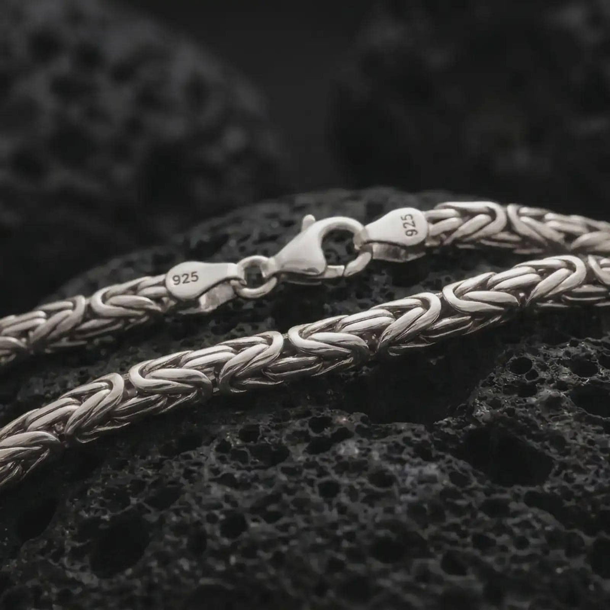 The polished silver 4mm round Byzantine chain necklace is displayed on textured black volcanic rock, highlighting the woven link pattern, lobster clasp, and 925 silver stamp in a premium macro photograph.