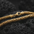 Load image into Gallery viewer, A gold plated 4mm round Byzantine necklace lies across rugged volcanic rock, featuring rounded interlocking links and a refined masculine chain profile in a luxury macro studio close-up.
