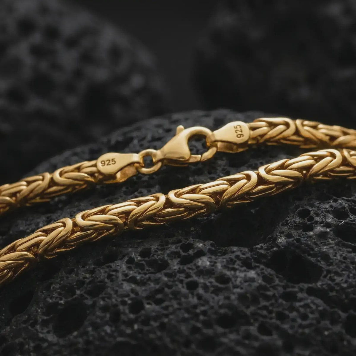 A gold plated 4mm round Byzantine necklace lies across rugged volcanic rock, featuring rounded interlocking links and a refined masculine chain profile in a luxury macro studio close-up.