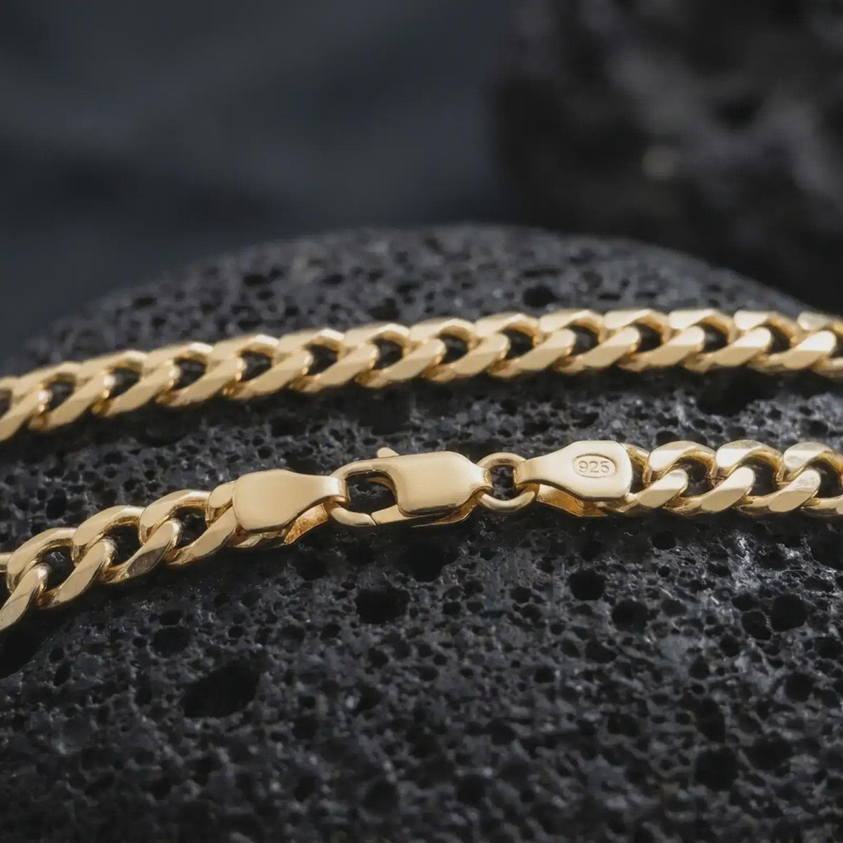 A gold plated 5mm Cuban chain necklace lies on rugged volcanic rock, featuring smooth flattened links and a refined masculine chain profile in a luxury macro studio close-up.