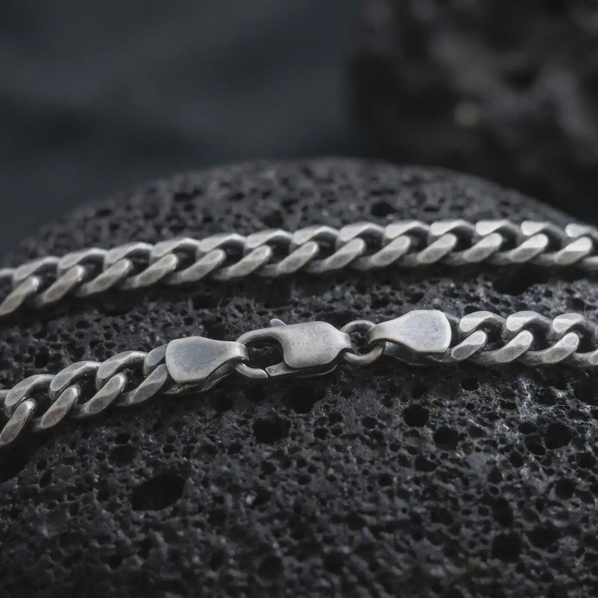A 5mm Cuban chain necklace with an oxidized finish rests on dark volcanic rock, showing its flattened curb links and classic sterling silver structure in a macro studio close-up.