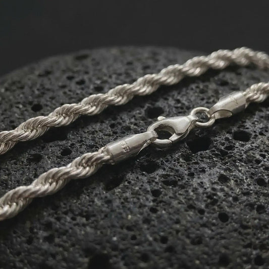 The polished silver 3mm rope chain necklace is displayed on textured black volcanic rock, highlighting the braided twist pattern, secure clasp, and refined everyday profile in a premium macro photograph.