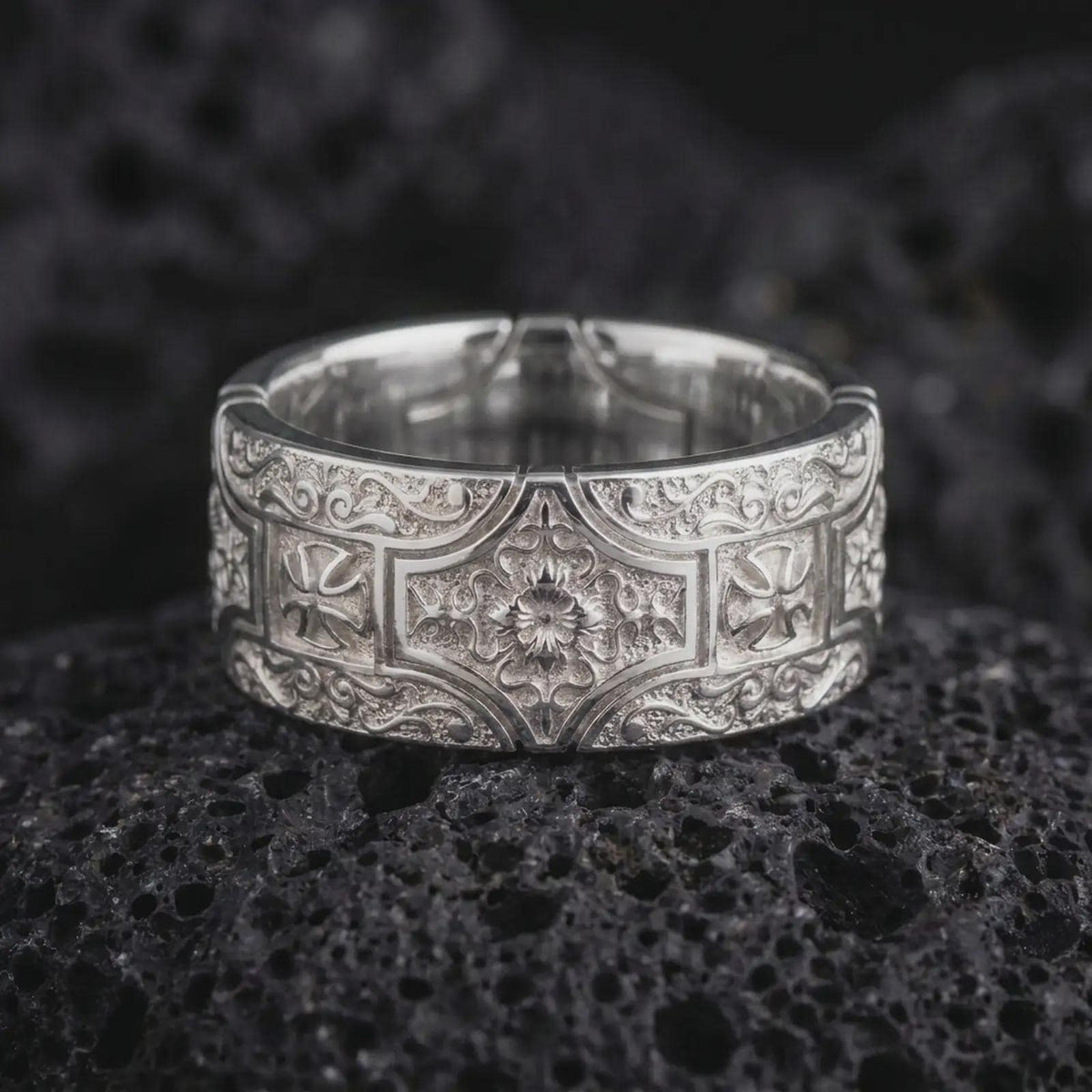 The polished silver ornate cross ring is displayed on textured black volcanic rock, highlighting its symmetrical ornamental design, raised silver details, smooth rounded edges, and wide band profile in a premium macro photograph.