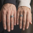 Load image into Gallery viewer, An oxidized sterling silver cross band ring is worn on a finger, displaying the dark recessed background, raised ornamental cross motifs, floral centerpiece, and substantial wide band profile in a close-up lifestyle photograph.
