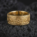 Load image into Gallery viewer, A gold plated ornate cross wedding band rests on rugged volcanic rock, showcasing raised cross motifs, flowing scrollwork, a detailed floral centerpiece, and dark recessed detailing in a luxury macro studio close-up.
