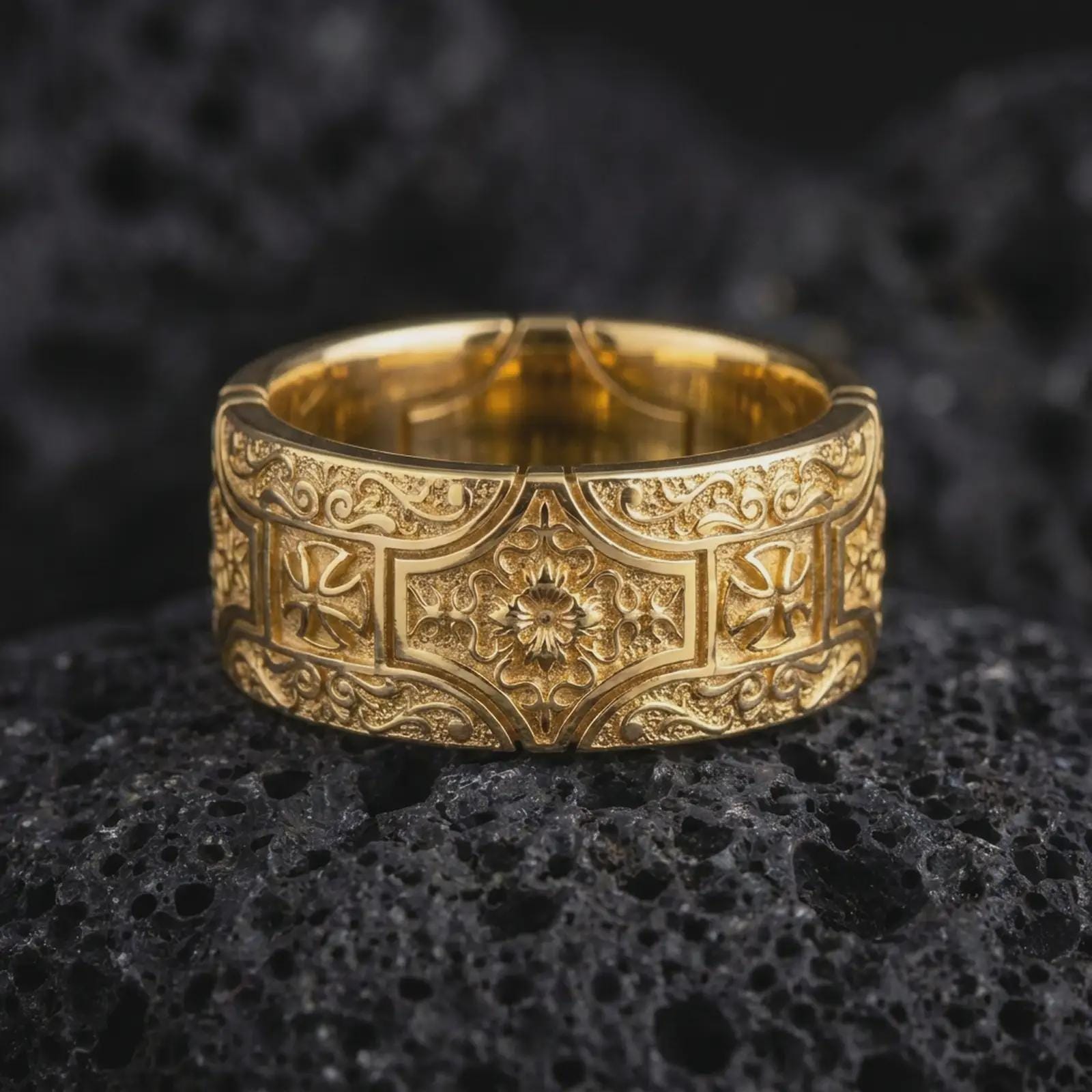 A gold plated ornate cross wedding band rests on rugged volcanic rock, showcasing raised cross motifs, flowing scrollwork, a detailed floral centerpiece, and dark recessed detailing in a luxury macro studio close-up.