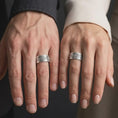 Load image into Gallery viewer, The polished silver ornate wedding band is worn naturally on a finger, showing the symmetrical cross-inspired pattern, detailed floral centerpiece, smooth rounded edges, and bold sterling silver band design in a premium lifestyle photograph.
