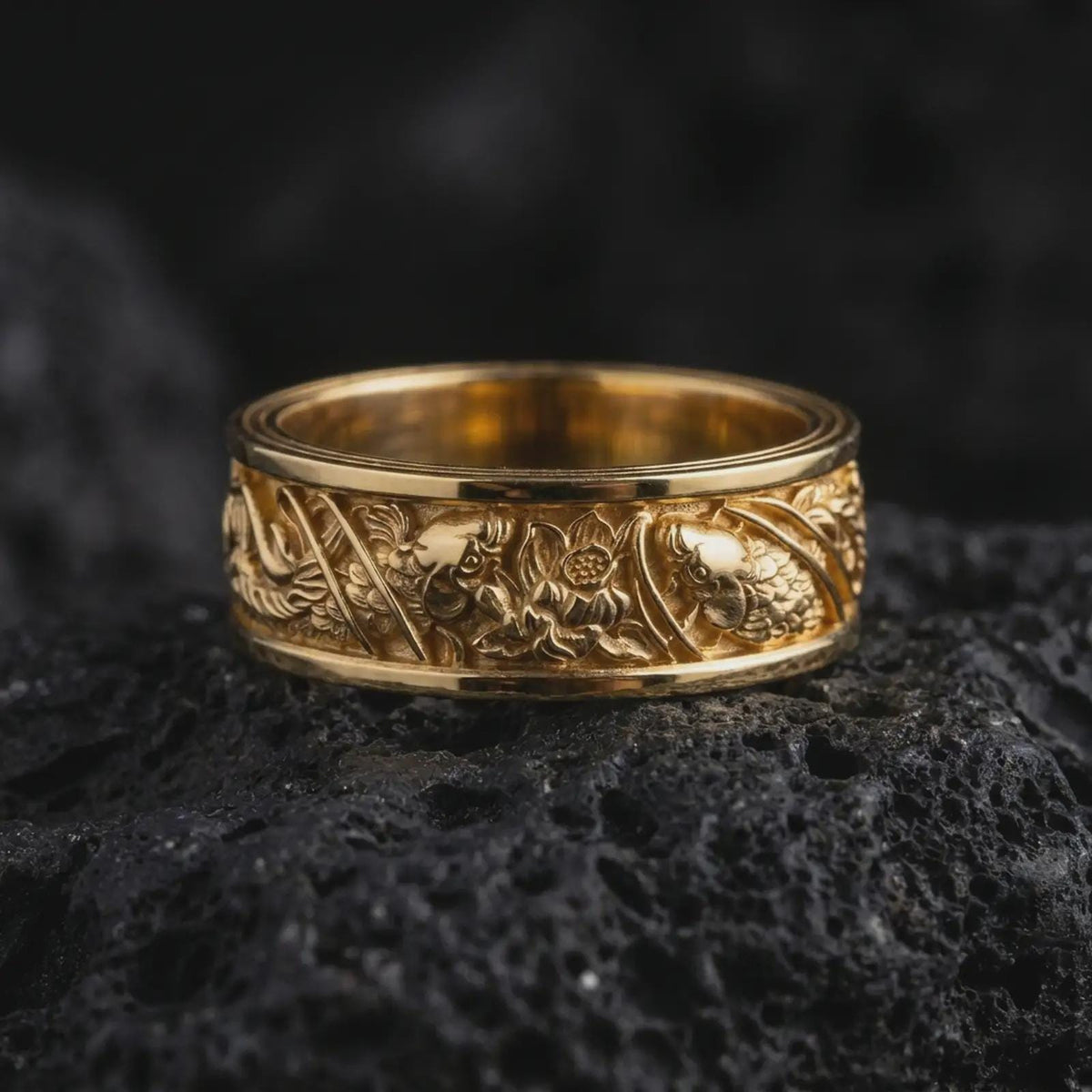 A gold plated floral silver ring rests on rugged volcanic rock, showcasing raised flowers, botanical foliage, dark recessed detailing, and an ornate wide band profile in a luxury macro studio close-up.