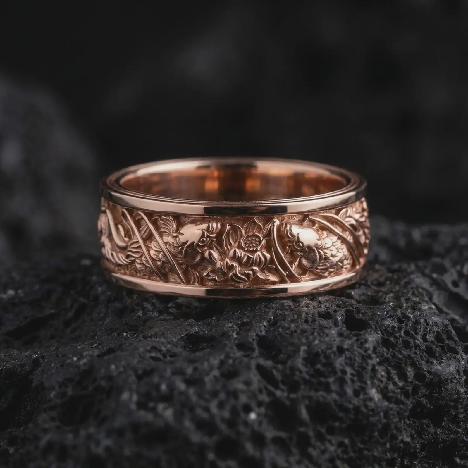 The rose gold plated botanical wedding band is arranged on black volcanic rock, emphasizing its detailed floral relief, flowing leaves, organic ornamentation, and elegant nature-inspired design.