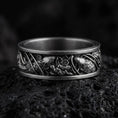 Load image into Gallery viewer, An oxidized sterling silver floral band ring rests on dark volcanic rock, featuring raised flowers, leaves, and intricate botanical details around the wide band in a macro studio close-up.
