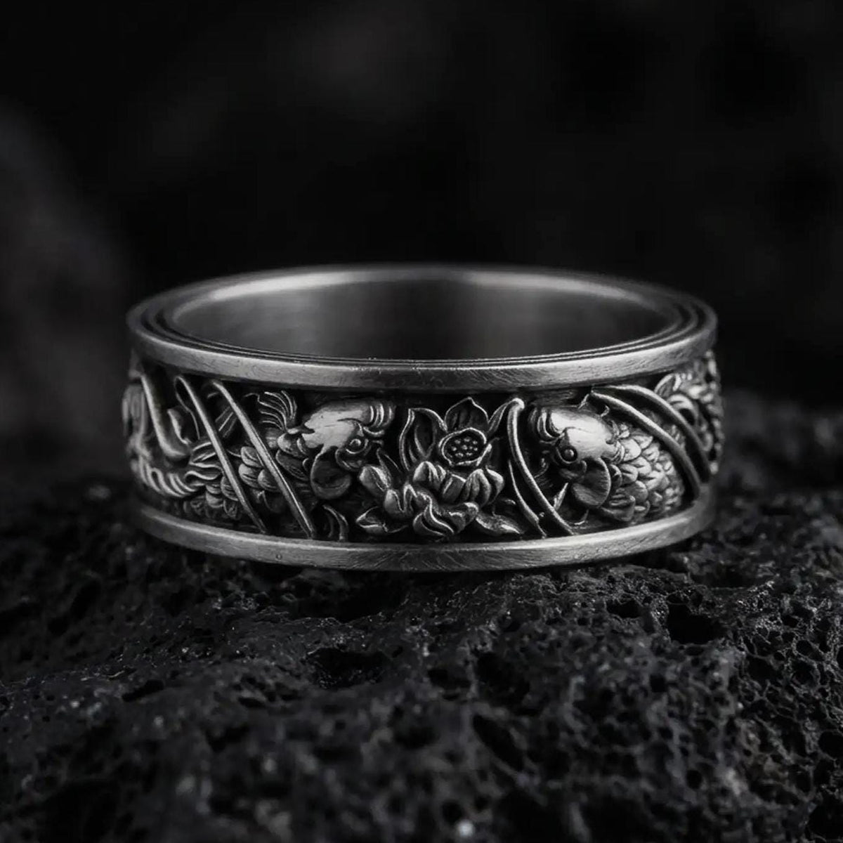 An oxidized sterling silver floral band ring rests on dark volcanic rock, featuring raised flowers, leaves, and intricate botanical details around the wide band in a macro studio close-up.