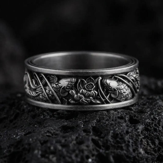 An oxidized sterling silver floral band ring rests on dark volcanic rock, featuring raised flowers, leaves, and intricate botanical details around the wide band in a macro studio close-up.