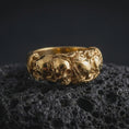 Load image into Gallery viewer, A gold plated sterling silver skull ring rests on rugged volcanic rock, showcasing a raised skull surrounded by sculpted roses and leaves, with detailed recessed textures creating strong visual depth in a luxury macro studio close-up.
