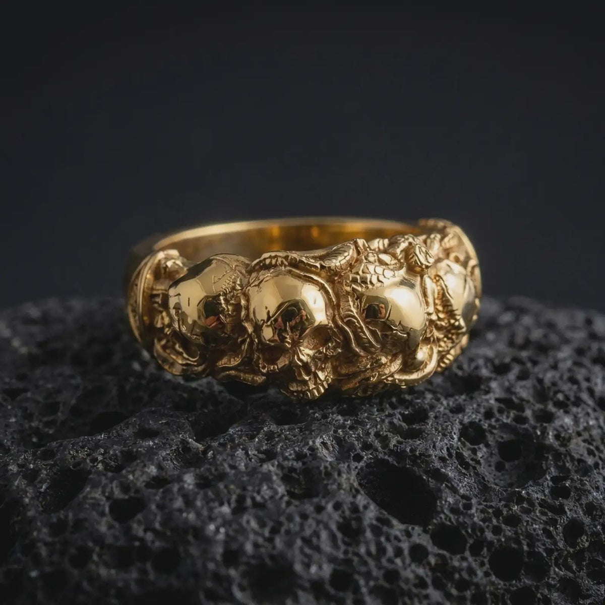 A gold plated sterling silver skull ring rests on rugged volcanic rock, showcasing a raised skull surrounded by sculpted roses and leaves, with detailed recessed textures creating strong visual depth in a luxury macro studio close-up.