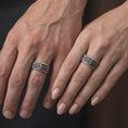 Load image into Gallery viewer, An oxidized Baroque fleur-de-lis spinner ring is worn on a finger, displaying dark recessed texture, raised silver scrollwork, and a bold wide band profile in a close-up lifestyle photograph.
