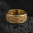 Load image into Gallery viewer, A gold plated Baroque spinner ring rests on rugged volcanic rock, featuring repeating fleur-de-lis motifs, elaborate scrollwork, and a wide textured wedding band profile in a luxury macro studio close-up.
