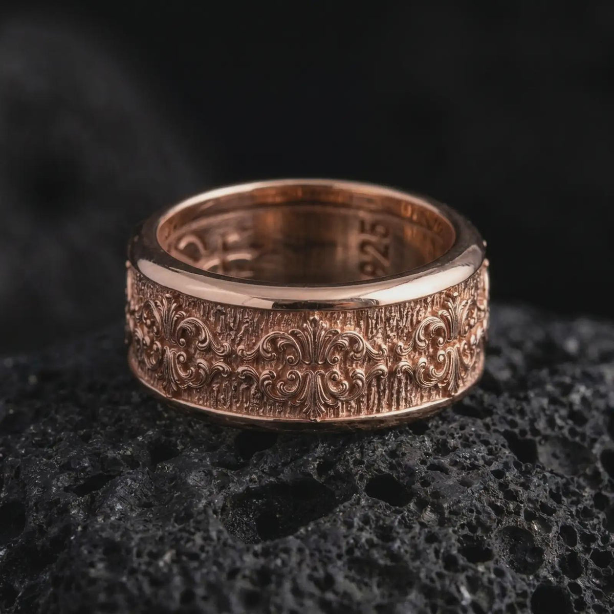 The rose gold plated fleur-de-lis wedding band is arranged on black volcanic rock, emphasizing the raised ornamental pattern, vintage-inspired texture, and refined unisex spinner ring construction in a detailed macro image.