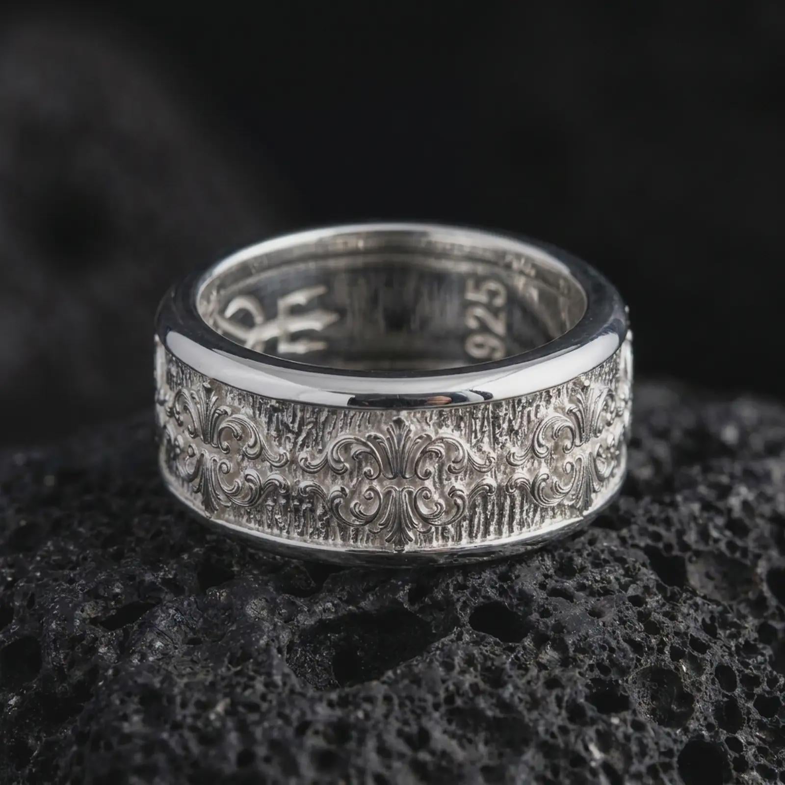 The polished silver fleur-de-lis spinner ring is displayed on textured black volcanic rock, highlighting the symmetrical Baroque scrollwork, raised ornamental details, and smooth rounded edges in a premium macro photograph.