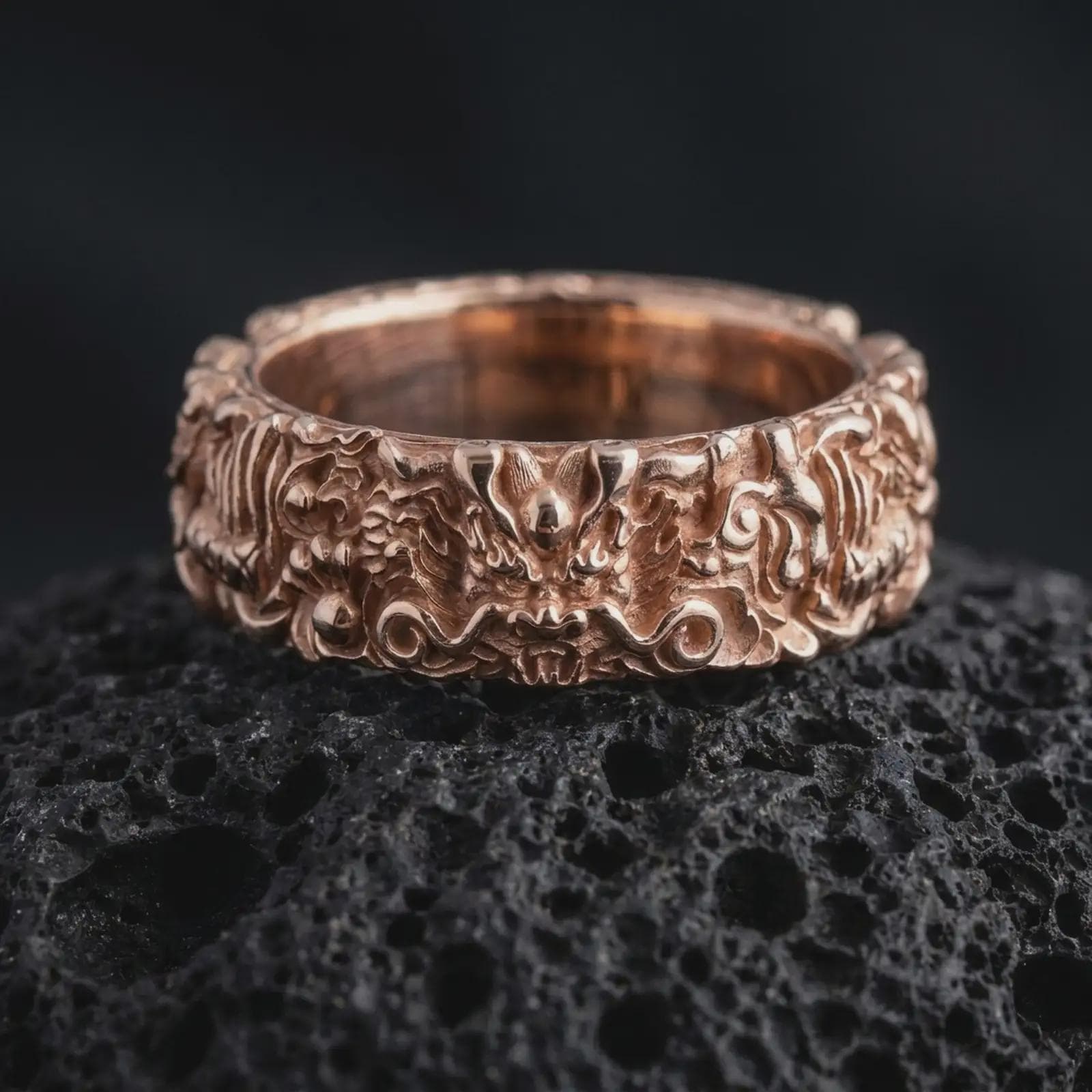 The rose gold plated dragon wedding ring is arranged on black volcanic rock, emphasizing its sculpted mythical face, ornamental surrounding details, deep relief work, and bold wide band construction in a refined macro image.