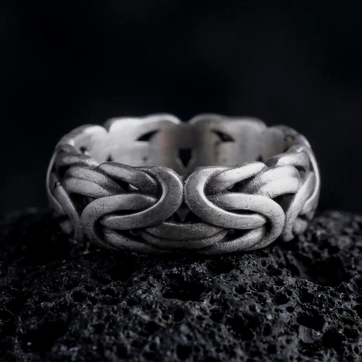 An oxidized sterling silver Byzantine chain band ring rests on dark volcanic rock, showing sculpted interlocking links, woven chain texture, and a substantial wide band in a macro studio close-up.