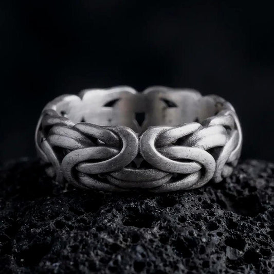 An oxidized sterling silver Byzantine chain band ring rests on dark volcanic rock, showing sculpted interlocking links, woven chain texture, and a substantial wide band in a macro studio close-up.