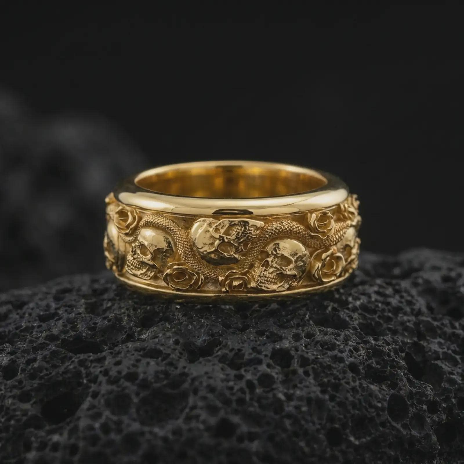 A gold plated skull snake ring rests on rugged volcanic rock, showcasing raised skull motifs, a coiling serpent, detailed roses, and dramatic memento mori-inspired relief in a luxury macro studio close-up.