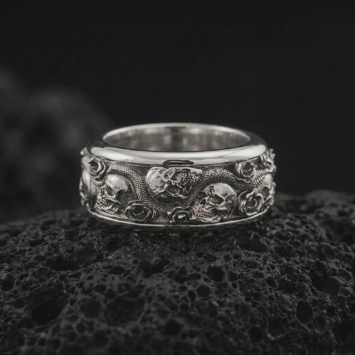 The polished silver gothic skull and snake ring is displayed on textured black volcanic rock, highlighting the three-dimensional skulls, serpent scales, floral roses, and substantial wide band profile in a premium macro photograph.