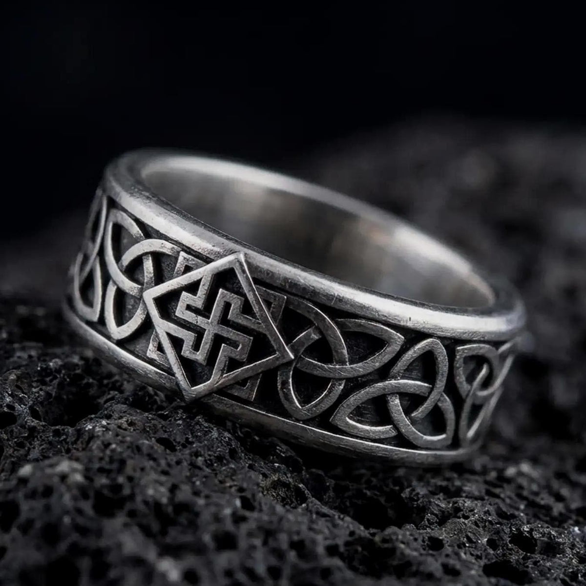 An oxidized sterling silver Celtic knot ring rests on dark volcanic rock, showing raised interlaced knotwork and a dark recessed band background in a macro studio close-up.