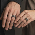 Load image into Gallery viewer, An oxidized floral spinner ring is worn on a finger, displaying the dark recessed background, raised silver flowers, and symbolic wide band design in a close-up lifestyle shot.

