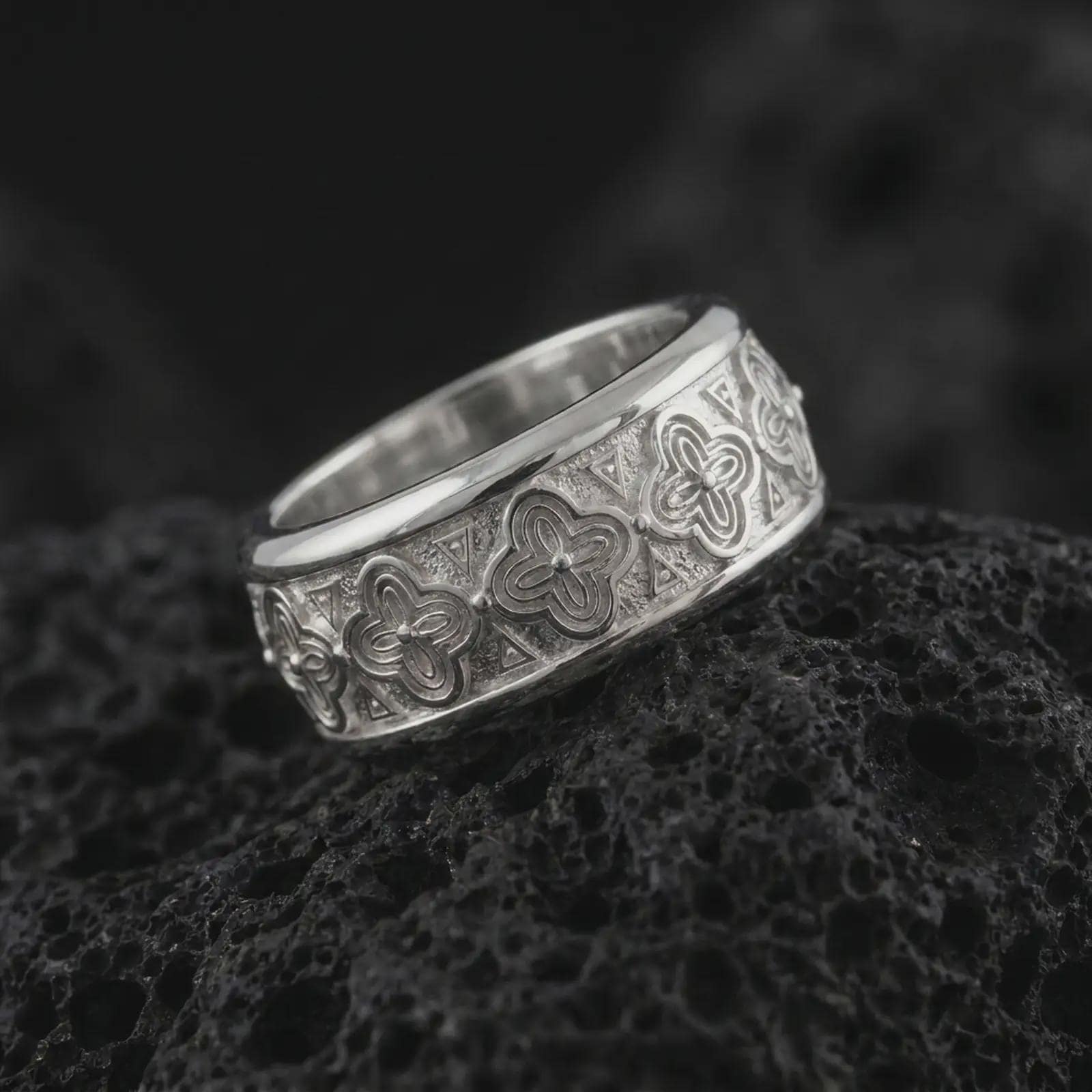 The polished silver flower wedding band is displayed on textured black volcanic rock, highlighting the repeating floral pattern, smooth rounded edges, and wide spinner ring profile in a premium macro photograph.