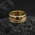 Load image into Gallery viewer, A gold plated Grand Teton mountain spinner ring sits on rugged volcanic rock, featuring an engraved mountain panorama and a refined wide band silhouette in a luxury macro close-up.
