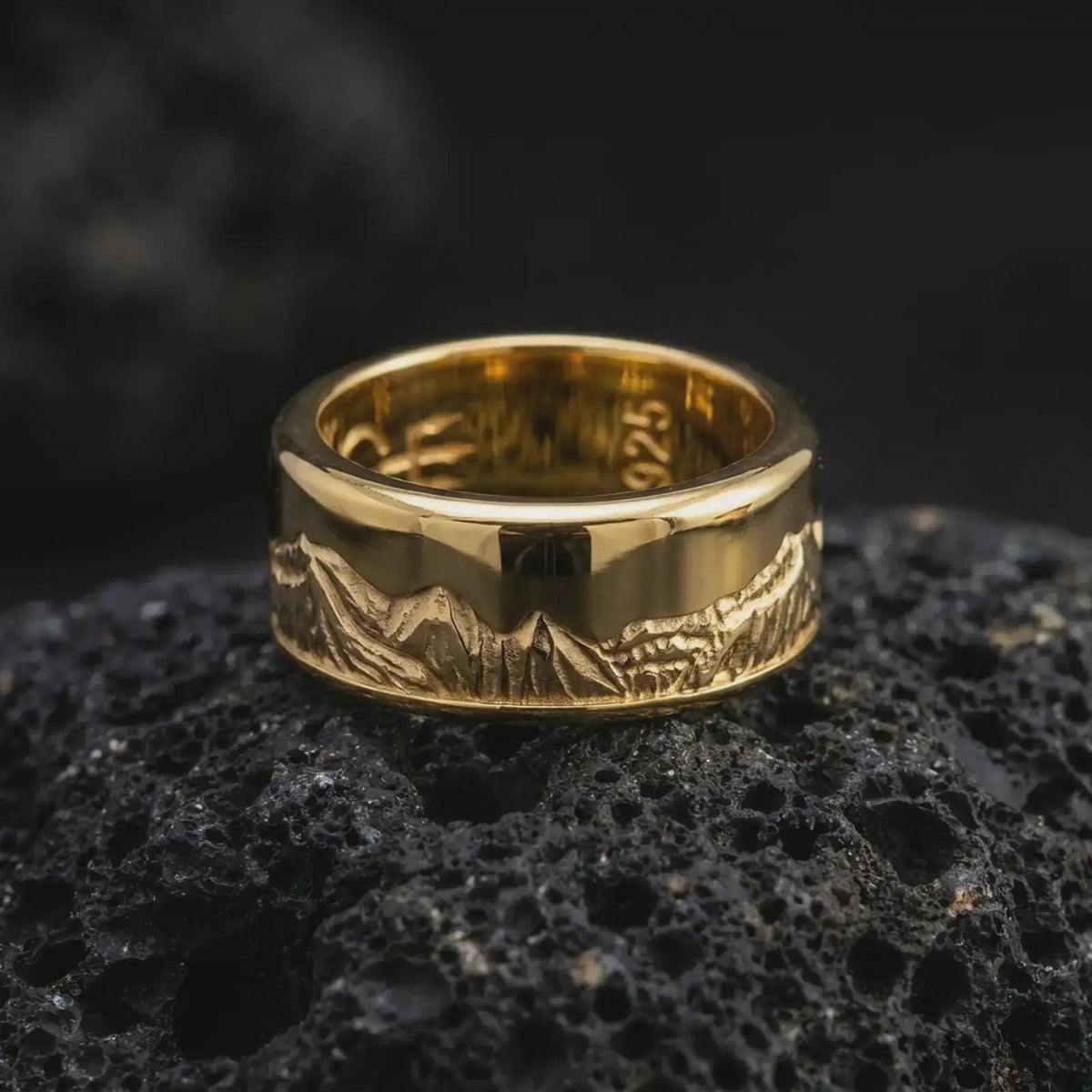 A gold plated Grand Teton mountain spinner ring sits on rugged volcanic rock, featuring an engraved mountain panorama and a refined wide band silhouette in a luxury macro close-up.