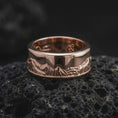 Load image into Gallery viewer, The rose gold plated mountain wedding band is presented on dark volcanic rock, emphasizing the engraved Grand Teton landscape and smooth spinning band profile in a detailed product image.
