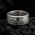 Load image into Gallery viewer, An oxidized sterling silver Grand Teton mountain spinner ring rests on dark volcanic rock, showcasing a detailed mountain landscape engraved around the wide band in a macro product photograph.
