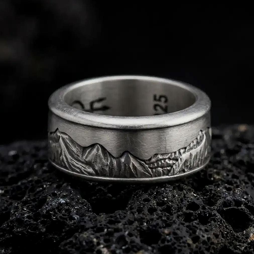 An oxidized sterling silver Grand Teton mountain spinner ring rests on dark volcanic rock, showcasing a detailed mountain landscape engraved around the wide band in a macro product photograph.