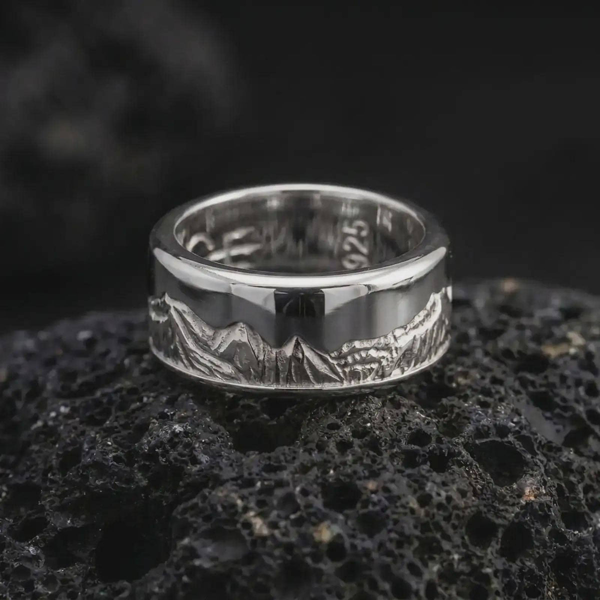 The polished silver Grand Teton mountain ring is displayed on black volcanic rock, highlighting the scenic mountain engraving and spinning fidget band design in a clean macro studio image.
