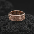 Load image into Gallery viewer, The rose gold plated floral meditation ring is arranged on black volcanic rock, emphasizing its botanical pattern, smooth rounded borders, and textured recessed background in a refined macro image.
