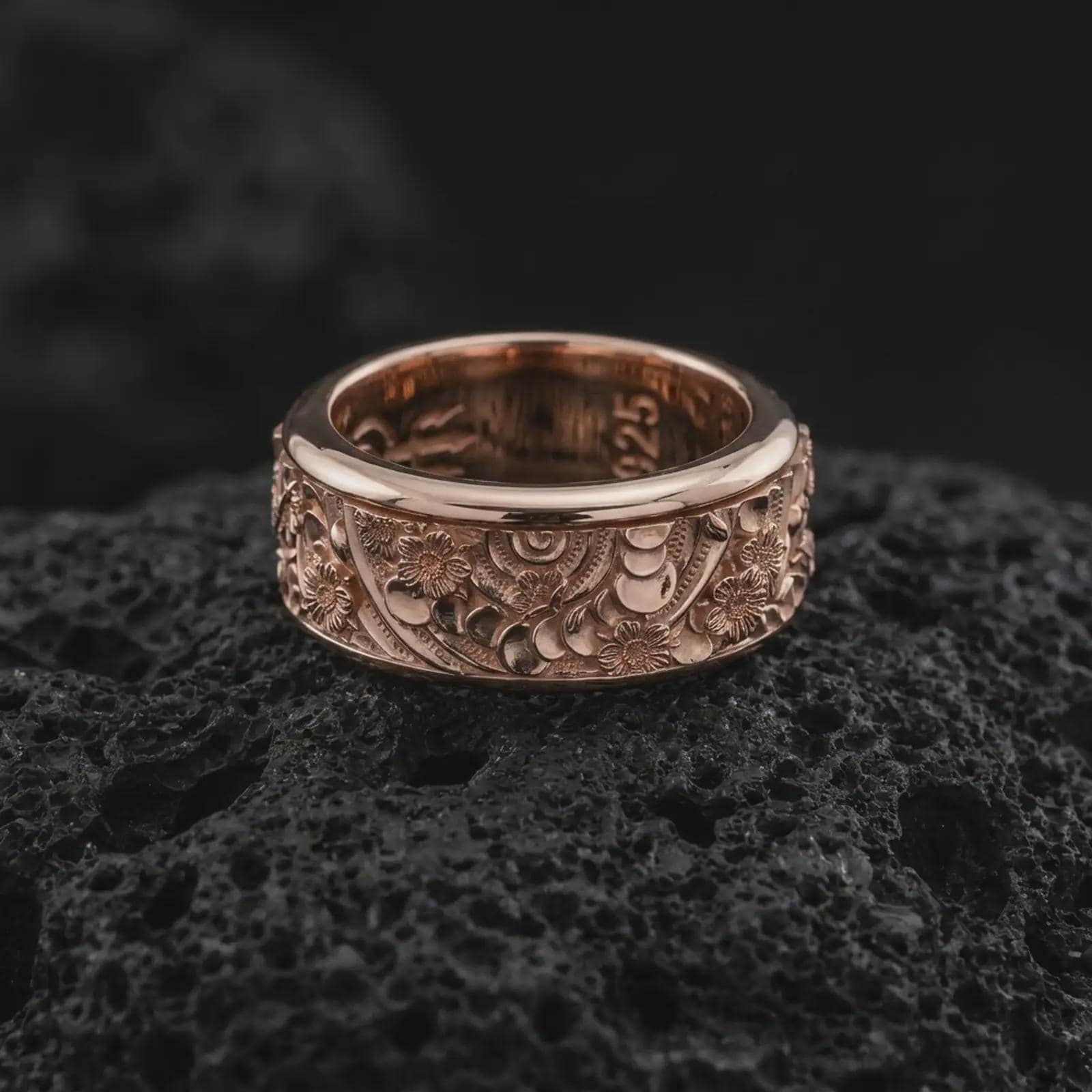 The rose gold plated floral meditation ring is arranged on black volcanic rock, emphasizing its botanical pattern, smooth rounded borders, and textured recessed background in a refined macro image.