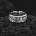 Load image into Gallery viewer, The polished silver floral spinner ring is displayed on textured black volcanic rock, highlighting the wide band, rounded edges, botanical engravings, and detailed flower relief in a premium macro photograph.
