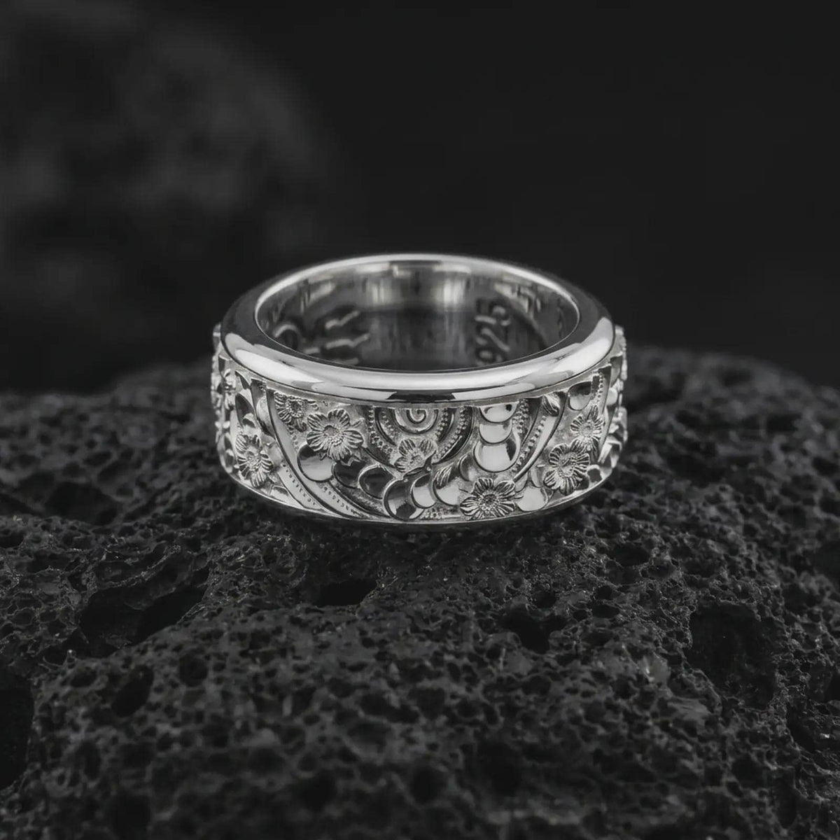 The polished silver floral spinner ring is displayed on textured black volcanic rock, highlighting the wide band, rounded edges, botanical engravings, and detailed flower relief in a premium macro photograph.