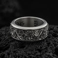 Load image into Gallery viewer, An oxidized sterling silver spring flower spinner ring rests on dark volcanic rock, showing raised floral blossoms, curved petal patterns, and dark recessed texture in a macro studio close-up.
